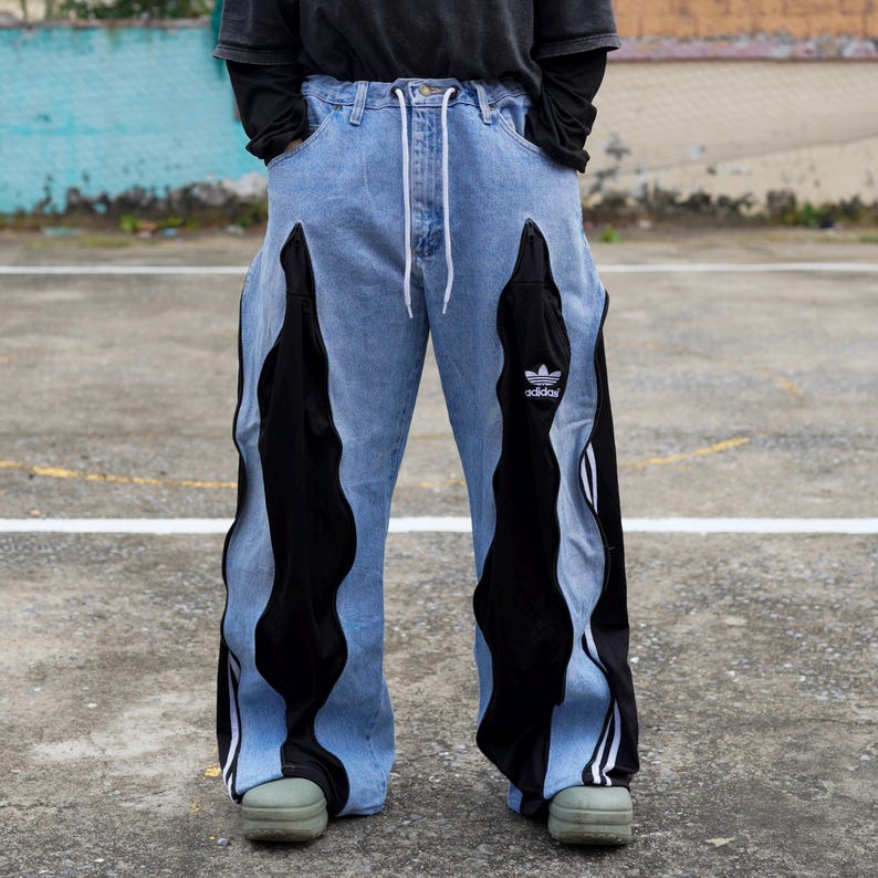 May include: Light blue denim jeans with black wavy panels down the sides. The jeans have a white drawstring, a small Adidas logo, and white stripes on the black panels. The pants are wide-legged and worn with green shoes.