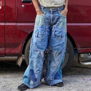 Op de afbeelding: Een persoon draagt ​​een blauwe jeans gemaakt van meerdere lagen denim, met zichtbare naden en pleisters. De jeans is erg wijd en heeft een ontspannen pasvorm.