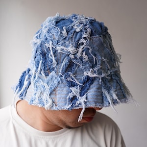 以下が含まれることがあります： 青いデニムのバケットハット。ダメージ加工と切りっぱなしのデザインが特徴です。フリンジ加工された縁とパッチワークのような外観が、ユニークで質感のある外観を作り出しています。ファッションアクセサリーです。