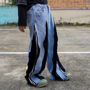 May include: Light blue denim pants with black and white accents. The pants feature vertical black panels with white stripes and a drawstring waist. The design includes a unique split leg detail and a relaxed fit.