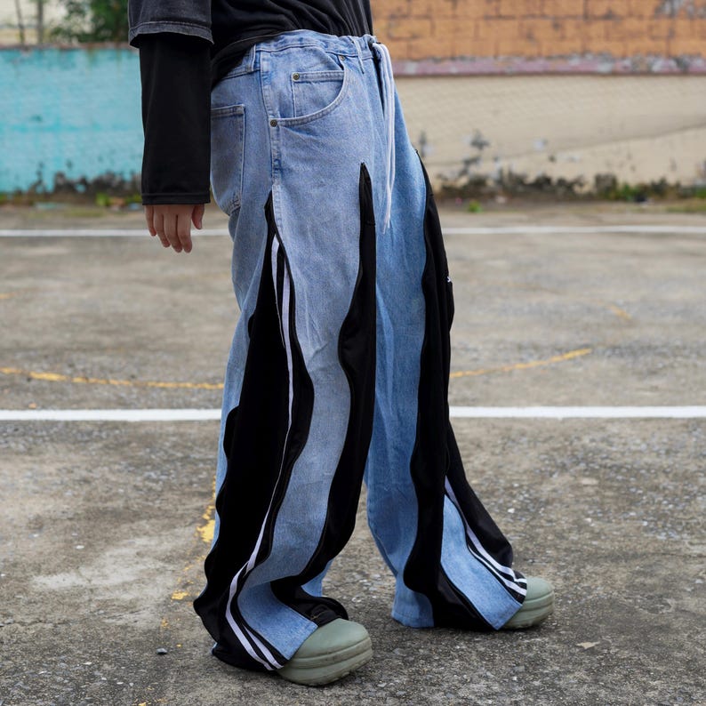 May include: Light blue denim pants with black and white accents. The pants feature vertical black panels with white stripes and a drawstring waist. The design includes a unique split leg detail and a relaxed fit.