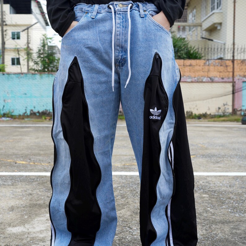 May include: Light blue denim jeans with black panels and white stripes. The jeans feature a drawstring waist and the Adidas logo on the black panel. The design incorporates a unique, wavy cut.