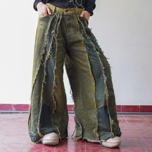 Op de afbeelding: Wijd uitlopende jeans in groen en blauw denim met een distressed, gelaagd ontwerp. De jeans hebben rafelige randen en een unieke patchwork-stijl, wat een textuurrijke, vintage look creëert. Het model draagt de jeans met een zwarte top en beige schoenen.