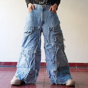 May include: Light blue denim jeans with a patchwork design. The jeans feature multiple layers of denim, pockets, and distressed details. The unique design creates a layered, textured appearance. The jeans are worn with white sneakers.