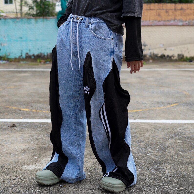 May include: Light blue denim jeans with black panels and white stripes. The jeans feature a drawstring waist and the Adidas logo. The lower legs have a unique design, and the person is wearing green shoes.