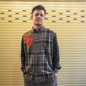 Könnte beinhalten: Ein kariertes Button-Down-Hemd mit gestickten roten Rosen auf der linken Seite. Das Hemd hat lange schwarze Ärmel und wird von einer Person getragen. Der Hintergrund ist eine gelbe Metalltür.