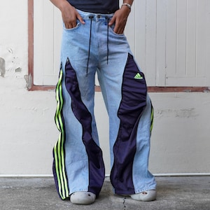 Puede incluir: Vaqueros de mezclilla azul claro con perneras anchas y paneles integrados en azul marino oscuro. El diseño incluye rayas laterales en verde neón brillante y un logotipo de Adidas en verde neón en el muslo izquierdo. El pantalón tiene cintura con cordón y cremalleras.