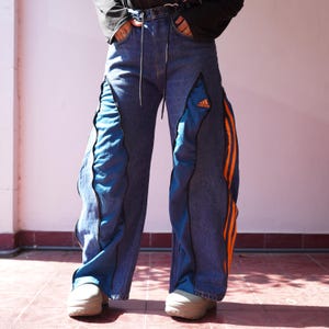 Peut inclure: Jean large en denim bleu avec des panneaux en tissu athlétique bleu intégrés sur le devant des jambes, des fermetures éclair noires et trois bandes verticales orange sur le côté extérieur. Le logo Adidas est brodé en orange sur le panneau avant.