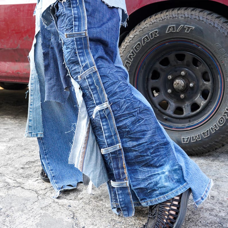 May include: A pair of blue denim jeans with multiple layers of fabric sewn together. The jeans have a distressed look and are ripped at the knees. The jeans are being worn by a person standing next to a vehicle.