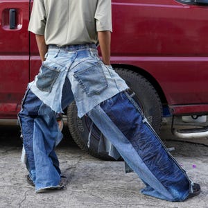 May include: A person wearing a pair of blue denim jeans that have been cut, layered, and sewn together to create a unique and stylish look. The jeans are distressed and have a patchwork design.