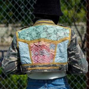 Könnte beinhalten: Eine Camouflage-Jeansjacke mit Patchwork-Design auf der Rückseite. Das Patchwork besteht aus verschiedenen Stoffen in Blautönen, Rosa und Gold. Die Jacke hat eine verkürzte Passform und ist mit goldenen Verzierungen verziert.
