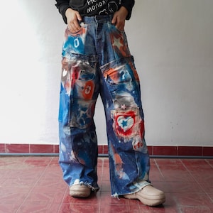 May include: Blue denim jeans with colorful patches and paint splatters. The jeans feature a patchwork design with various shapes and numbers. The jeans have a wide leg and a distressed hem. The jeans are worn with beige shoes.