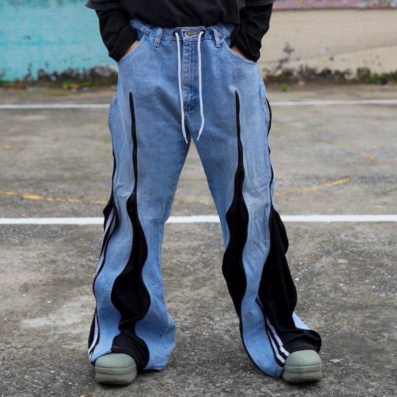 May include: Light blue denim jeans with black flame-like accents running down the legs. The jeans have a white drawstring and are paired with green shoes. The design features a unique, eye-catching style.