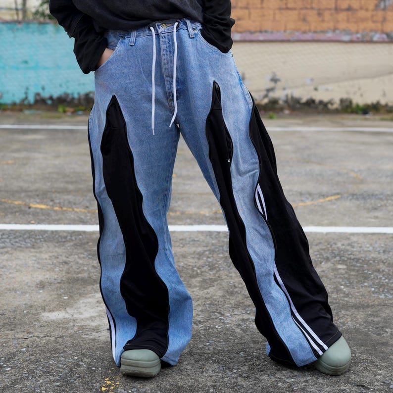 May include: Light blue denim pants with black fabric panels and white drawstring. The pants have a unique design with black fabric inserts that create a wavy pattern down the legs. The pants are wide-legged and have a casual style.