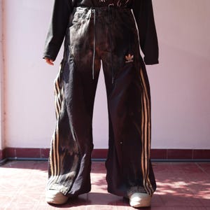 Op de afbeelding: Zwarte broek met wijde pijpen, een taille met trekkoord, een wit Adidas-trefoil-logo op de linkerdij en verticale zijpanelen met drie metallic gouden strepen. De stof heeft over het hele kledingstuk een vervaagde, verwassen uitstraling.