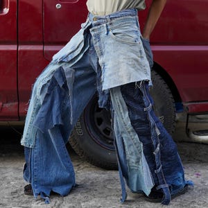 May include: A pair of blue denim jeans with multiple layers of fabric sewn together. The jeans are distressed and have a frayed hem. The jeans are being worn by a person standing next to a red truck.