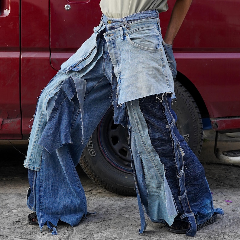 May include: A pair of blue denim jeans with multiple layers of fabric sewn together. The jeans are distressed and have a frayed hem. The jeans are being worn by a person standing next to a red truck.