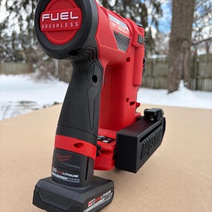 May include: A red and black cordless nail gun with the text "FUEL BRUSHLESS" on the top. The nail gun has a black handle and a black battery pack. The brand name "Milwaukee" is visible on the side.
