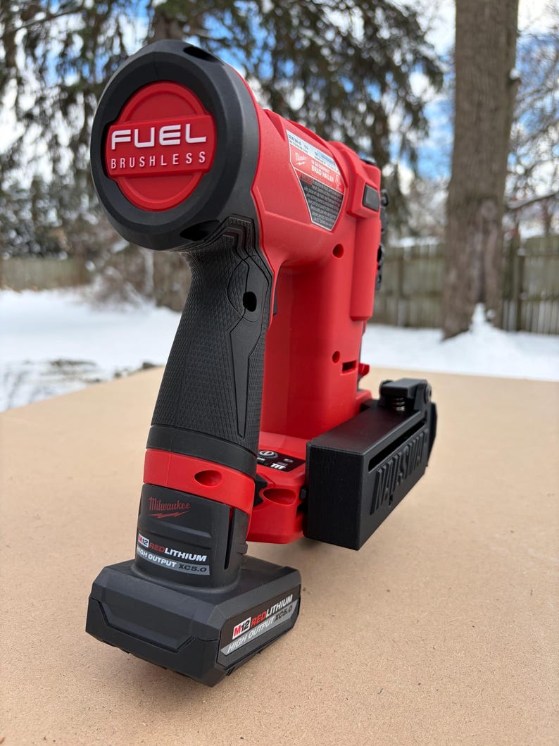 May include: A red and black cordless nail gun with the text "FUEL BRUSHLESS" on the top. The nail gun has a black handle and a black battery pack. The brand name "Milwaukee" is visible on the side.