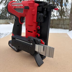 Op de afbeelding: Een rood-zwarte draadloze brad nailer met een zwarte "NAILSWAP"-bevestiging. De nailer staat op een bruin oppervlak met een besneeuwde achtergrond. Het heeft een batterij en een magazijn met spijkers.
