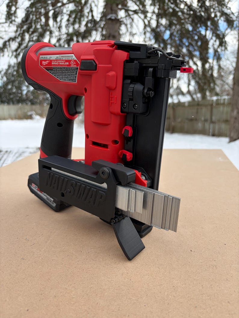 May include: A red and black cordless brad nailer with a black "NAILSWAP" attachment. The nailer is on a brown surface with a snowy background. The nailer has a battery and a magazine of nails.