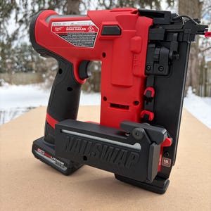 May include: A red and black Milwaukee 18-gauge Brad Nailer. The nailer has a black handle and a black magazine that reads "NAILSWAP". The nailer is sitting on a light brown surface.