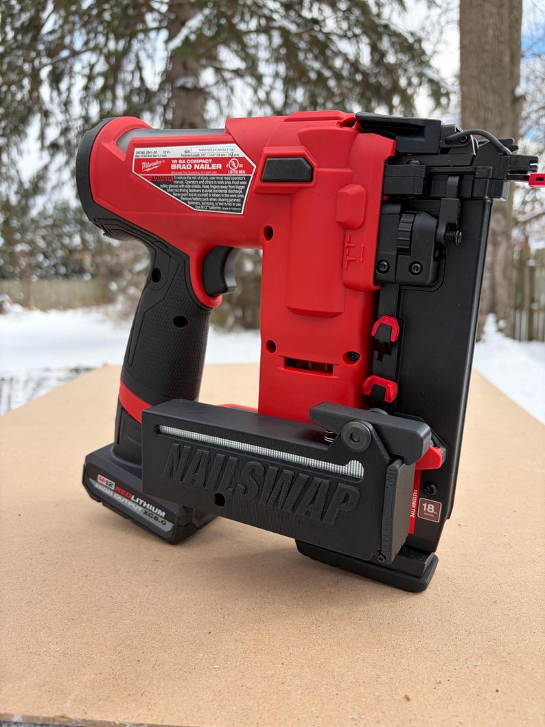 May include: A red and black Milwaukee 18-gauge Brad Nailer. The nailer has a black handle and a black magazine that reads "NAILSWAP". The nailer is sitting on a light brown surface.