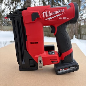 May include: A red and black Milwaukee FUEL cordless nail gun. The tool has a black grip, a black magazine, and the Milwaukee logo in white. The battery pack is black and has the text "M12 REDLITHIUM HIGH OUTPUT XC5.0".