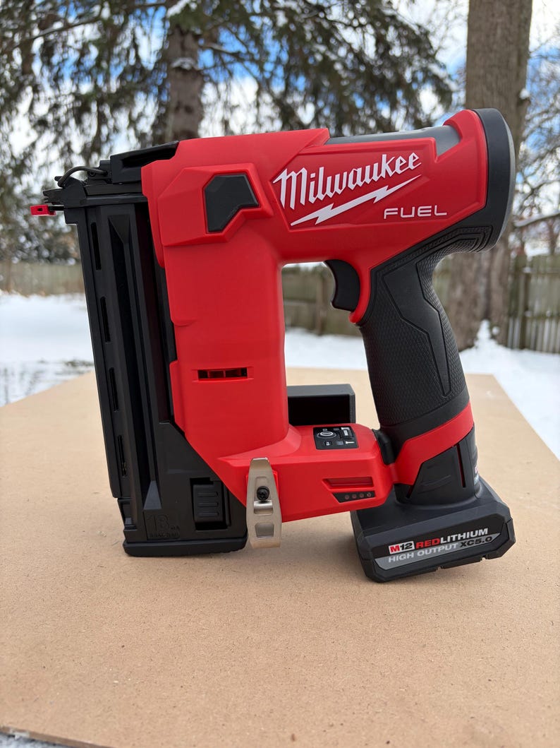 May include: A red and black Milwaukee FUEL cordless nail gun. The tool has a black grip, a black magazine, and the Milwaukee logo in white. The battery pack is black and has the text "M12 REDLITHIUM HIGH OUTPUT XC5.0".