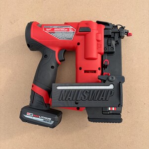 May include: A red and black cordless brad nailer with the text "NAILSWAP" on the front. The nailer has a black handle and a battery pack that reads "M12 REDLITHIUM HIGH OUTPUT XC5.0". The tool is on a light brown surface.