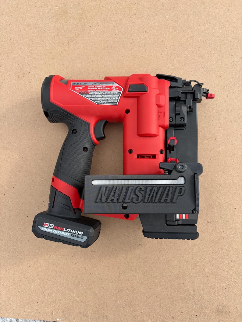 May include: A red and black cordless brad nailer with the text "NAILSWAP" on the front. The nailer has a black handle and a battery pack that reads "M12 REDLITHIUM HIGH OUTPUT XC5.0". The tool is on a light brown surface.