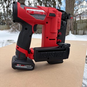 May include: A red and black cordless brad nailer with the text "NAILSWAP" on the front. The nailer has a black handle and a battery pack attached. The nailer is sitting on a brown surface.