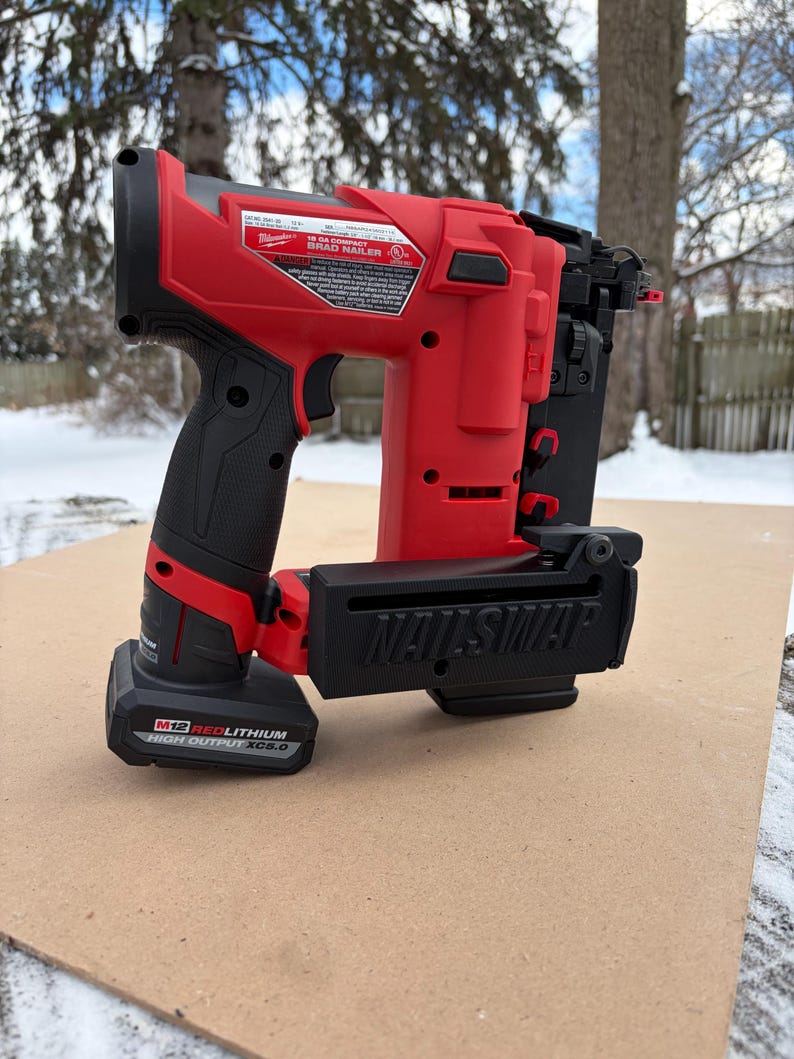 May include: A red and black cordless brad nailer with the text "NAILSWAP" on the front. The nailer has a black handle and a battery pack attached. The nailer is sitting on a brown surface.