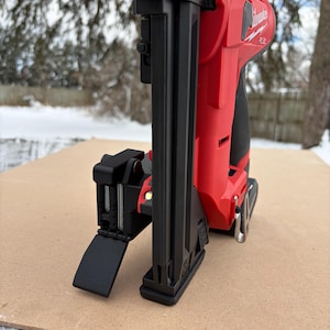 May include: A red and black cordless Milwaukee nail gun, angled slightly to the left, resting on a brown surface. The nail gun is set against a snowy backdrop with trees. The Milwaukee logo is visible on the side.