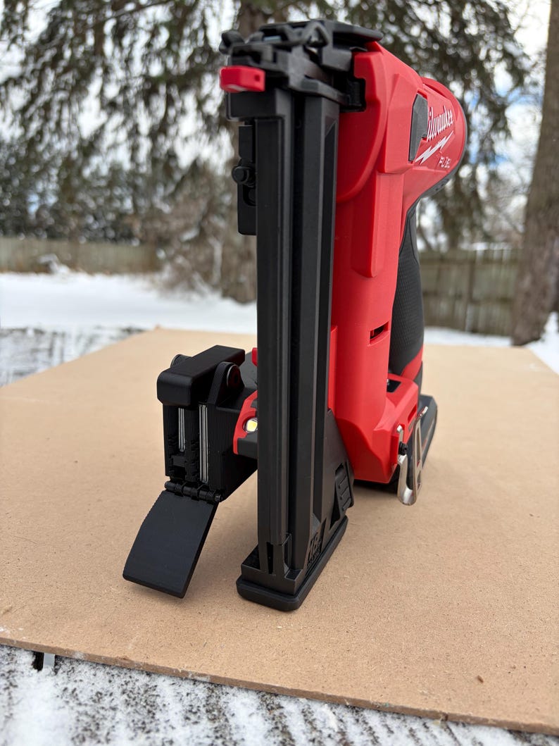 May include: A red and black cordless Milwaukee nail gun, angled slightly to the left, resting on a brown surface. The nail gun is set against a snowy backdrop with trees. The Milwaukee logo is visible on the side.