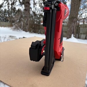 May include: A red and black cordless nail gun, resting on a brown surface, with a snowy outdoor background. The nail gun features a black nose, a red body, and a silver metal piece. The tool is designed for construction and woodworking projects.