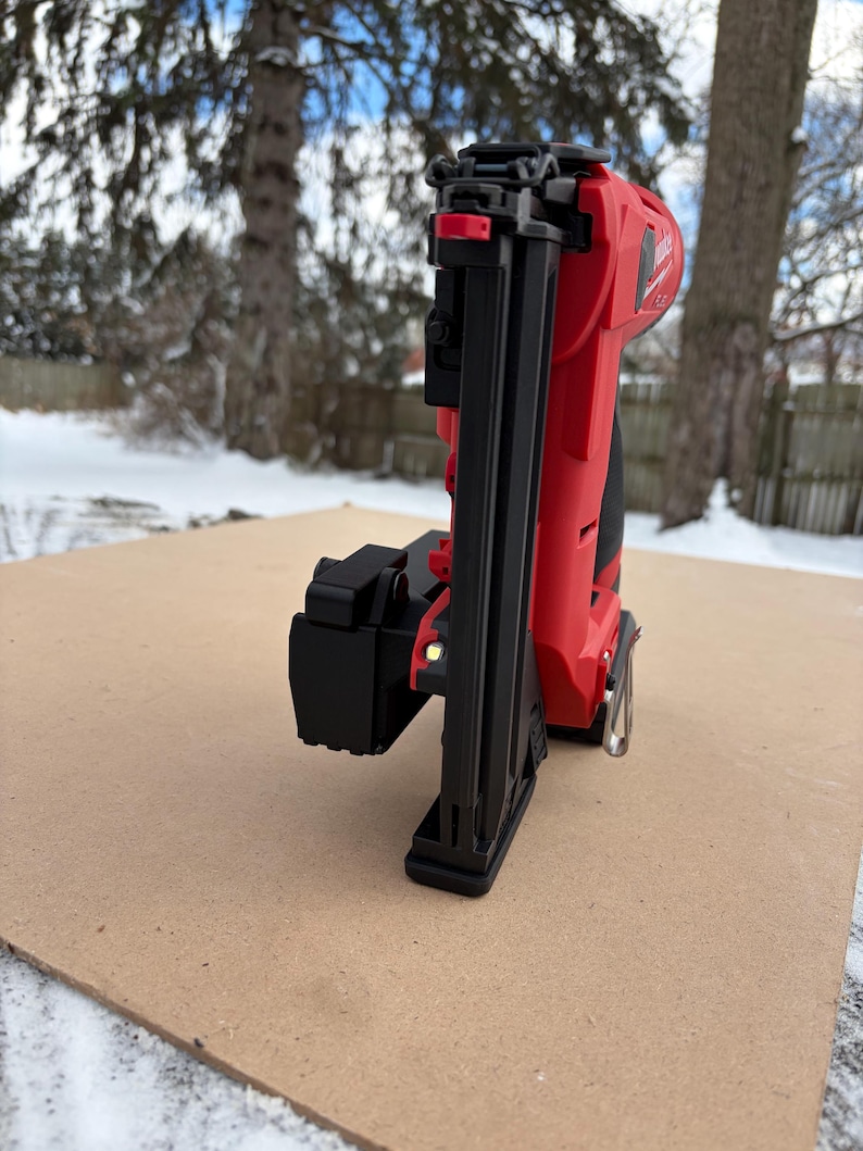 May include: A red and black cordless nail gun, resting on a brown surface, with a snowy outdoor background. The nail gun features a black nose, a red body, and a silver metal piece. The tool is designed for construction and woodworking projects.