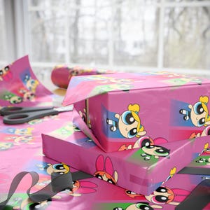 May include: Pink gift boxes wrapped in Powerpuff Girls-themed wrapping paper. The paper features the cartoon characters in various poses. A pair of scissors and a roll of matching wrapping paper are also visible.