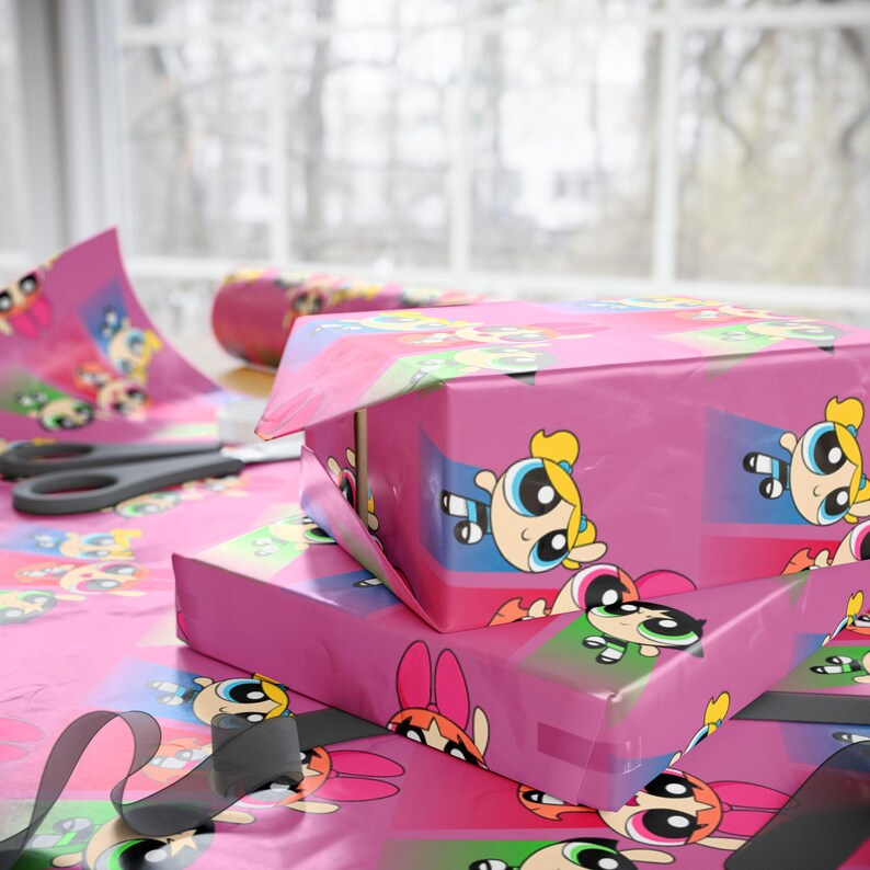 May include: Pink gift boxes wrapped in Powerpuff Girls-themed wrapping paper. The paper features the cartoon characters in various poses. A pair of scissors and a roll of matching wrapping paper are also visible.