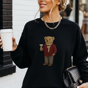 May include: Black sweatshirt featuring a cartoon bear in a red blazer, scarf, and pants, holding a martini glass. The bear is light brown with a white scarf. The sweatshirt is worn by a person holding a white coffee cup and a black purse.