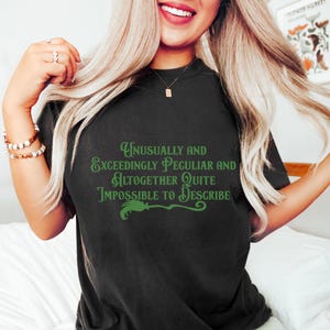 May include: Black t-shirt with green text that reads "Unusually and Exceedingly Peculiar and Altogether Quite Impossible to Describe." A green broom graphic is below the text. The shirt is a dark color.