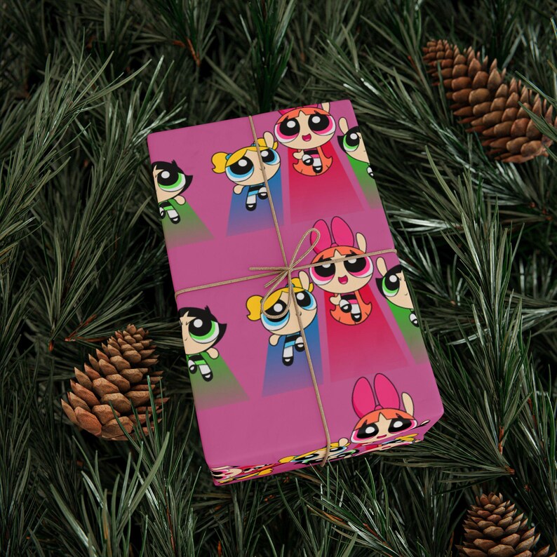 May include: A wrapped gift featuring a magenta background with a repeating pattern of cartoon characters in green, blue, and red. The gift is tied with twine and rests on a bed of green pine needles and pine cones.