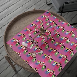 May include: A wrapped gift box and wrapping paper featuring a repeating pattern of cartoon characters on a pink background. The gift box is tied with a brown ribbon. A pair of scissors and a spool of ribbon are also visible.