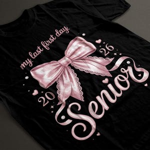 Peut inclure: T-shirt noir avec un nœud rose et les mots "my last first day" et "Senior 2026" en écriture cursive. Le nœud est orné d'une bordure en dentelle et entouré de cœurs et d'étoiles.