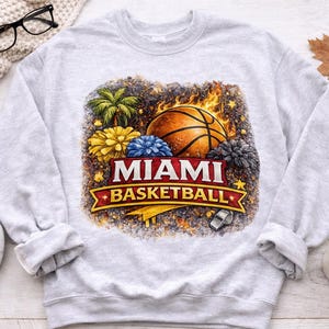 May include: A light gray sweatshirt featuring a basketball design with the words "MIAMI BASKETBALL" in red and white. The design includes a basketball, palm trees, and flowers. A whistle is also included in the design.