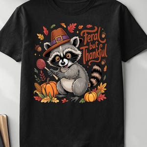 Peut inclure: T-shirt noir avec un raton laveur de dessin animé portant un chapeau marron et tenant une sucette. Le raton laveur est entouré de citrouilles, de feuilles et du texte "Feral but Thankful". Un t-shirt graphique amusant sur le thème de l'automne.