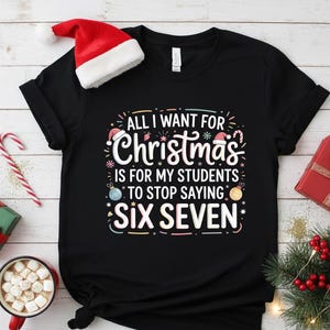 Peut inclure: T-shirt noir avec le texte "ALL I WANT FOR Christmas IS FOR MY STUDENTS TO STOP SAYING SIX SEVEN" dans un design festif. Un bonnet de Père Noël est posé sur le dessus. Des articles sur le thème de Noël entourent le t-shirt.