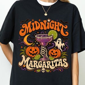 Pode incluir: Uma t-shirt preta com uma estampa gráfica com o texto Midnight Margaritas. O design inclui uma mão de esqueleto segurando um copo de margarita roxo, duas abóboras laranja, um fantasma branco, uma lua crescente amarela e morcegos.