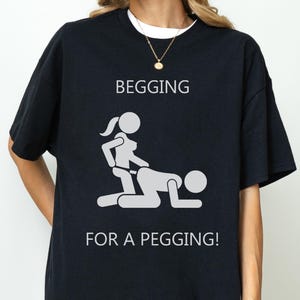May include: A black short-sleeve t-shirt featuring a white graphic of two stick figures in a sexual position. The text above the graphic reads BEGGING and the text below the graphic reads FOR A PEGGING! The shirt has a crew neckline.