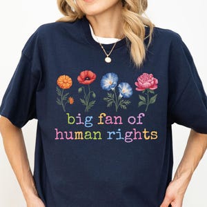 Könnte beinhalten: Marineblaues T-Shirt mit floralem Design und dem Text "big fan of human rights" in bunten Buchstaben. Das Shirt hat einen Rundhalsausschnitt und kurze Ärmel. Das florale Design umfasst orange, rote, blaue und rosa Blumen.